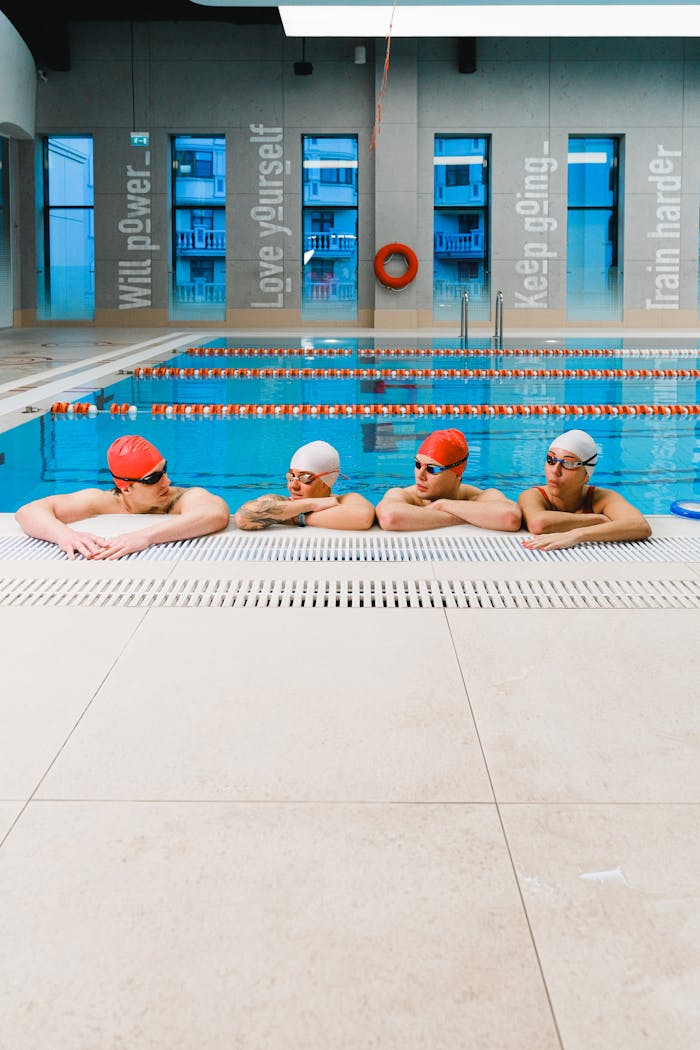 Four adult swimmers in red and white caps resting at an indoor swimming pool, preparing for training.